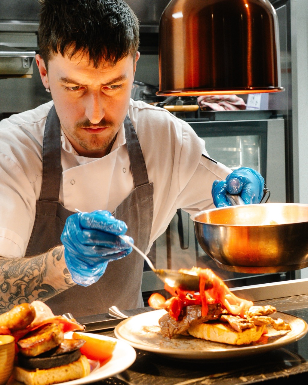 http://Chef%20in%20a%20white%20uniform%20and%20blue%20gloves%20plates%20a%20stacked%20burger%20with%20peppers%20in%20a%20professional%20kitchen;%20stainless%20bowl%20nearby.