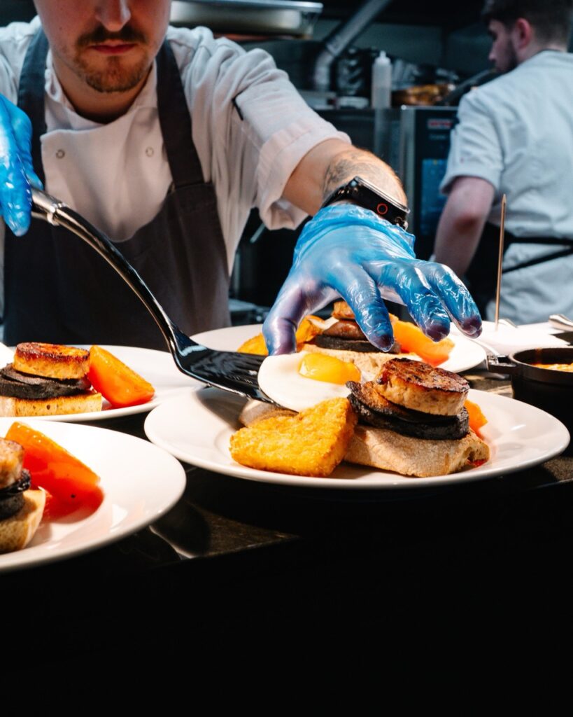 Chef in a white uniform and blue gloves plates multiple dishes with toast, seared meat, and vegetables in a bustling kitchen.