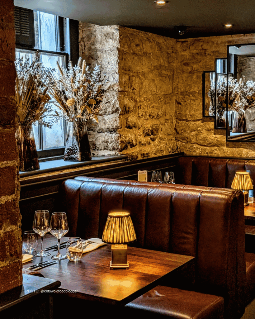 Cozy dining nook with brown leather booths, stone walls, a lit table lamp, and a table set with wine glasses.