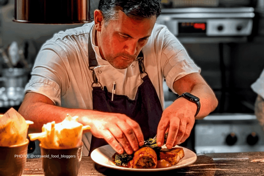 Male chef in a white shirt and dark apron plates a dish at a wooden counter in a professional kitchen.