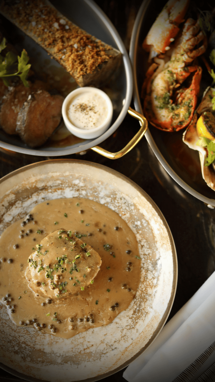 Beef in a creamy peppercorn sauce garnished with herbs on a white plate, with bread and a seafood dish in the background.